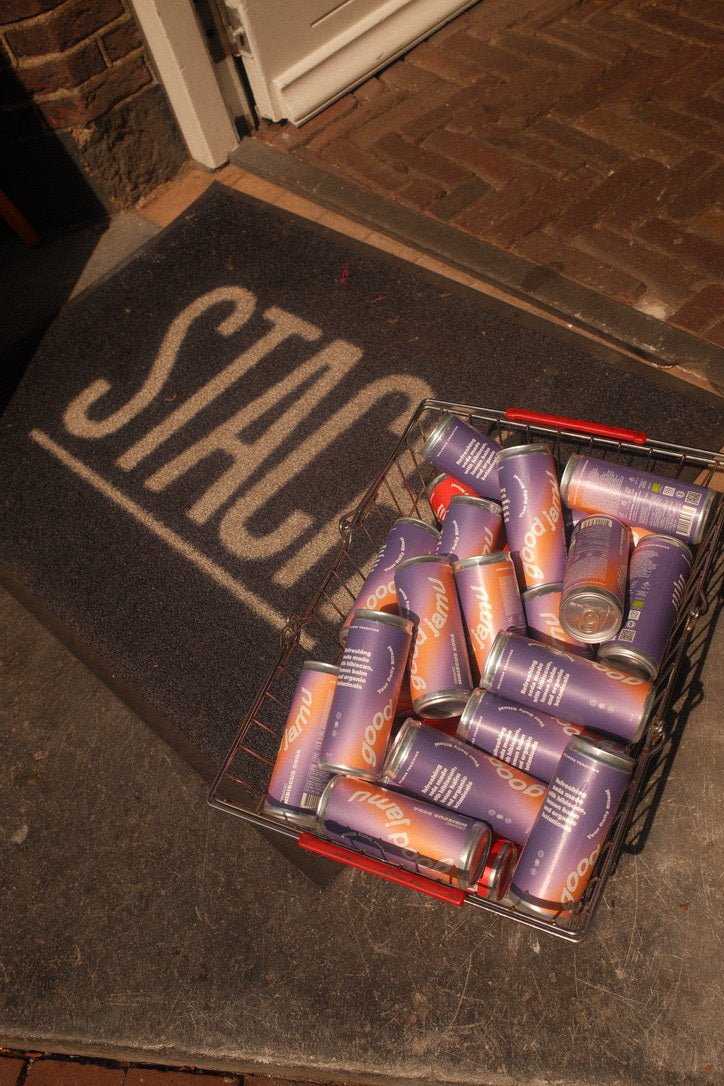 A wire basket filled with purple and orange cans of Good Jamu soda placed on the doorstep of Stach store, with the Stach logo visible on the entrance mat in warm, natural sunlight