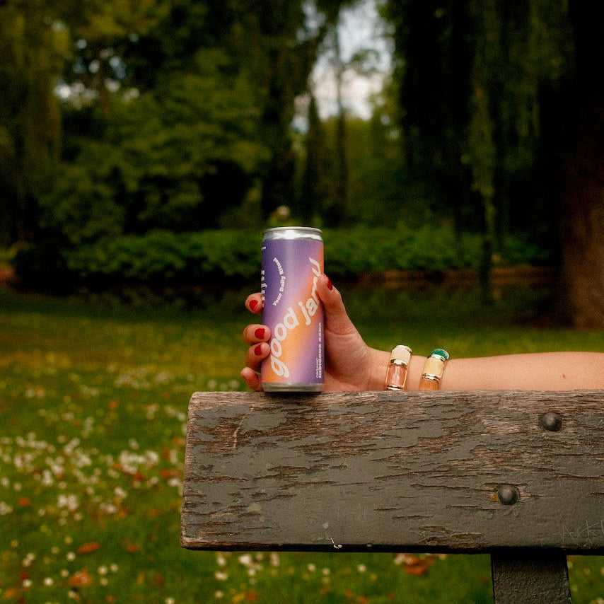 Hand houdt paarse Good Jamu soda uit de Starterpack vast op parkbank. Perfecte verfrissing voor buitenmomenten met natuurlijke ingrediënten.